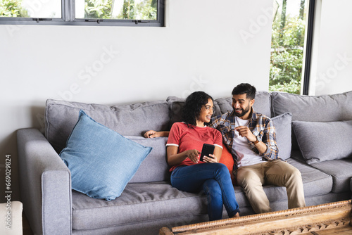 Happy biracial couple sitting on sofa and using smartphone in living room at home photo