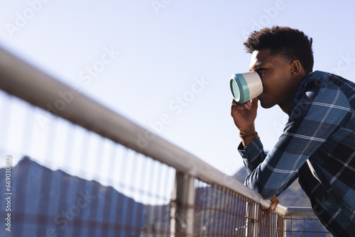 African american man drinking coffee standing on sunny balcony, copy space