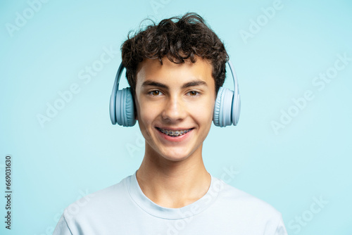 Portrait of attractive smiling boy with dental braces wearing wireless headphones isolated on blue background. Technology, advertisement concept 