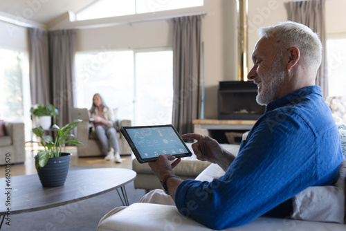 Happy senior caucasian man sitting on sofa using tablet with smart home interface