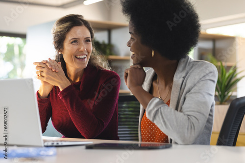 Happy diverse female colleagues discussing work at table with laptop in office photo
