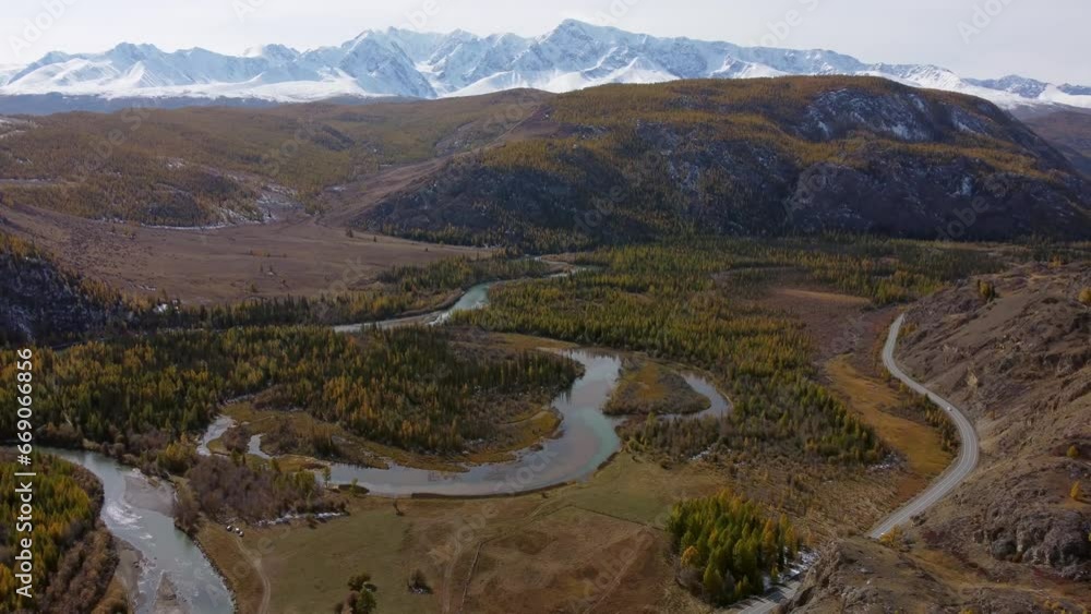 Slow motion aerial view of green tree hills with meandering flowing river water and motorable roadway in Altai, Russia against snow capped mountains cloudless blue sky in daylight Slow motion aerial view of green tree hills with meandering flowing river water and motorable roadway in Altai, Russia against snow capped mountains cloudless blue sky in daylight