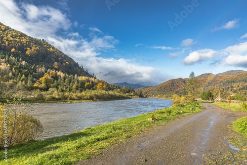 Autumn mountain landscape. Green tourism. Rural life. Orange and yellow trees in sunlight. Coniferous trees. Forest and mountains on the river bank. Country road along mountain river and forest.