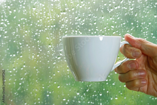 Hand holding a cup of coffee on window's glass with raindrops in the backdrop