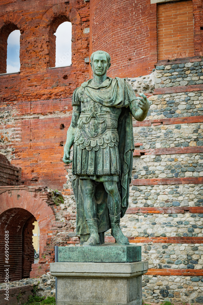 Naklejka premium Turin, Italy. Bronze statue depicting Julius Caesar, in front of the remains of the ancient Porta Palatina, datin gback to Roman times. Vertical image. 2023-05-04.