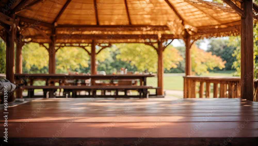 A wooden gazebo with a thatched roof in a park. The gazebo has a wooden table and benches. In the background is a blurred view of the park with trees and grass