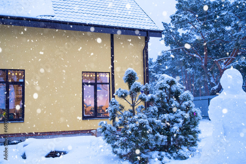 Cozy festive window of the house outside with the warm light of fairy lights garlands inside - celebrate Christmas and New Year in a warm home. Christmas tree, bokeh, snow on pine trees and snowfall © Ольга Симонова