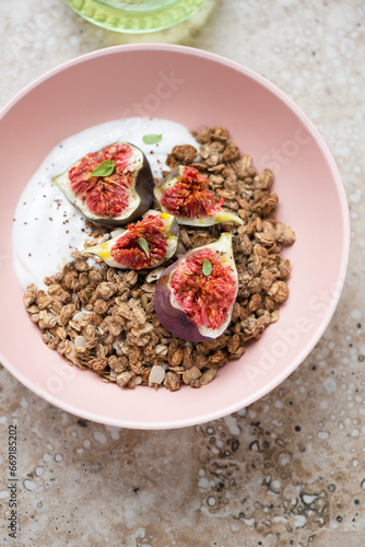 Granola with greek yogurt and torn fig fruits in a roseate bowl, vertical shot on a beige granite background, elevated view