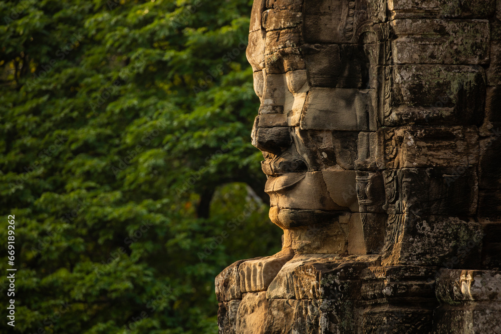 A huge face sculpted in stone, watches over visitors to the temple of Bayon, Angkor, Cambodia, in the background, a wall of green jungle surrounds it - obrazy, fototapety, plakaty 