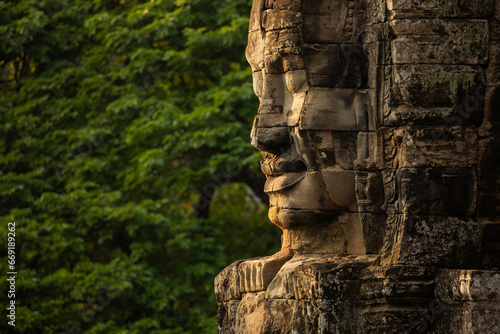 A huge face sculpted in stone, watches over visitors to the temple of Bayon, Angkor, Cambodia, in the background, a wall of green jungle surrounds it photo
