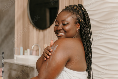Fototapeta Beautiful plus size African woman covered in towel applying hydrating cream on s