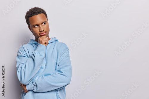 Horizontal shot of handsome curly haired African man keeps hand under chin concentrated aside pensively thinks over offer wears casual blue sweatshirt poses over white background. Let me think concept