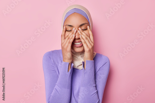 Horizontal shot of positive Muslim woman keeps eyes closed keeps hands on face smiles gladfully wears traditional hijab laughs at something funny isolated on pink background. Positive emotions concept photo