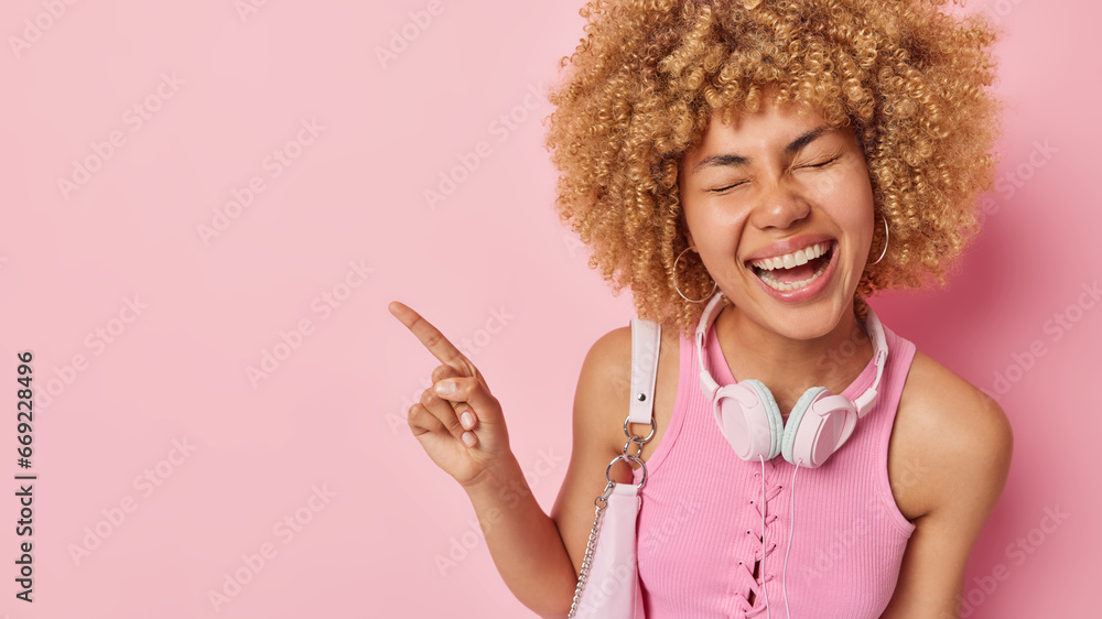 Overjoyed curly haired woman laughs gladfully shows something very funny indicates index finger on empty space dressed in casual t shirt with headphones around neck isolated over pink background