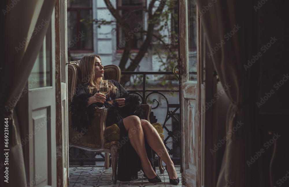 Fototapeta premium Lady in peignoir with a glass of wine. Sensual photo of European Blonde woman in black silk robe with feathers sleeve. Fashionable concept