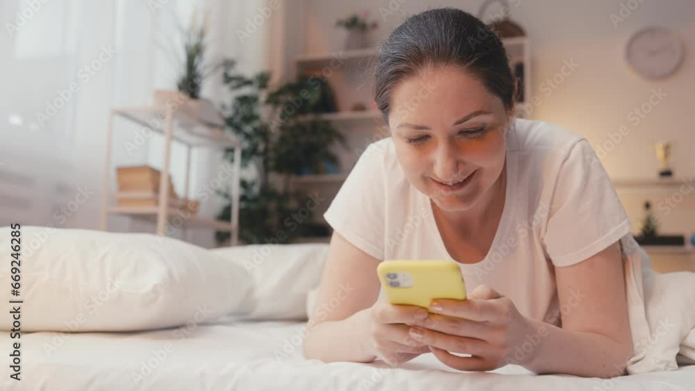Happy young woman resting on bed and texting in social networks on cellphone Happy young woman resting on bed and texting in social networks on cellphone