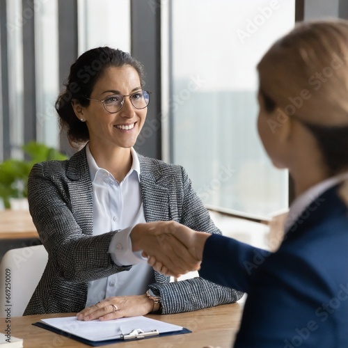 Mujer de mediana edad sonriente estrechando la mano de otra mujer en una reunión, con un contrato en la mesa.