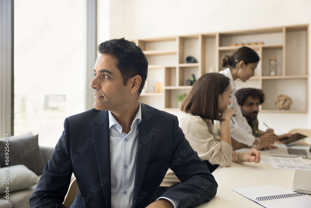 Thoughtful middle aged Indian businessman, CEO, company owner candid portrait. Pensive inspired business man looking away, sitting at meeting table with team cooperating in blurred background Thoughtful middle aged Indian businessman, CEO, company owner candid portrait. Pensive inspired business man looking away, sitting at meeting table with team cooperating in blurred background
