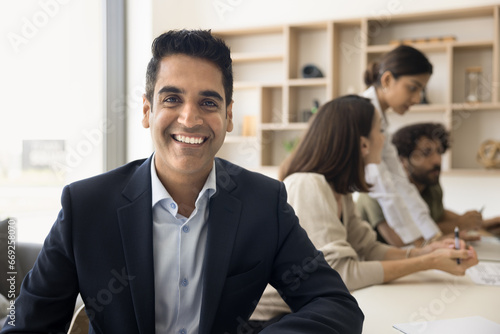 Cheerful handsome mature Indian business leader man professional portrait. Happy businessman, boss, company owner, project manager sitting at meeting table with team of employees working in background