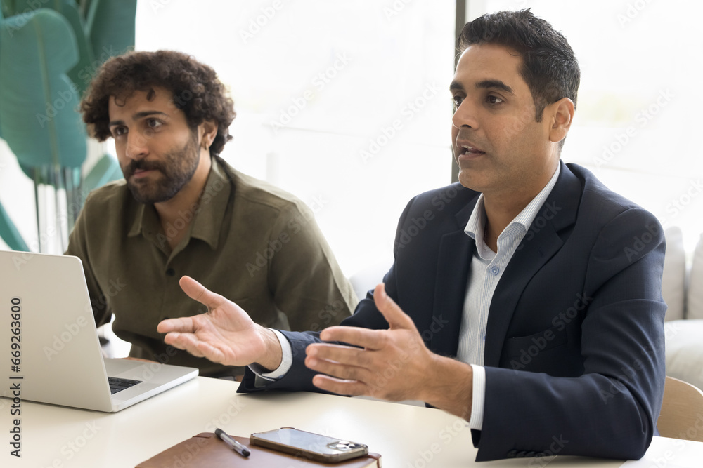 Serious Indian businessman in formal jacket talking to partners or colleagues on negotiation meeting at table. Diverse business colleagues discussing cooperation, project management, teamwork Serious Indian businessman in formal jacket talking to partners or colleagues on negotiation meeting at table. Diverse business colleagues discussing cooperation, project management, teamwork