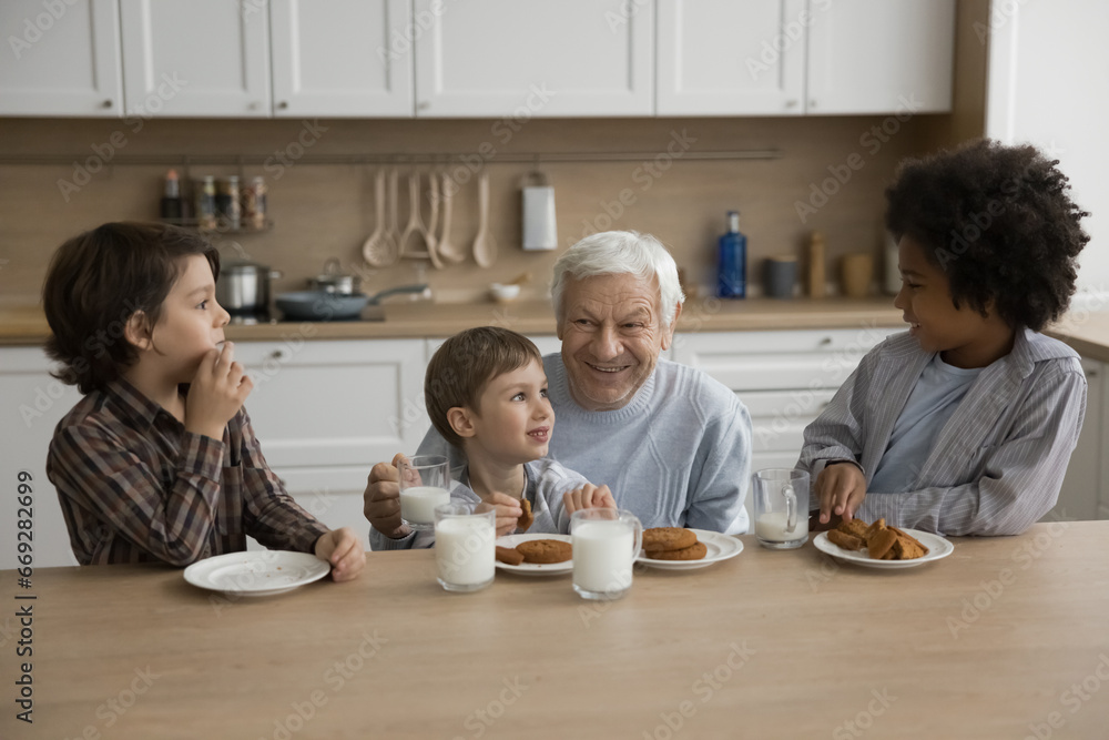 Multigenerational family, three cute great-grandchildren spend time with loving elderly great-grandpa sit in kitchen drinking milk eating delicious sweet homemade cookies, enjoy food and conversation Multigenerational family, three cute great-grandchildren spend time with loving elderly great-grandpa sit in kitchen drinking milk eating delicious sweet homemade cookies, enjoy food and conversation