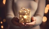 Happy woman holding Christmas gift with golden bow on background of christmas tree with lights. Stylish multiethnic lovers holding present close up in festive decorated room. Happy Holidays, at home