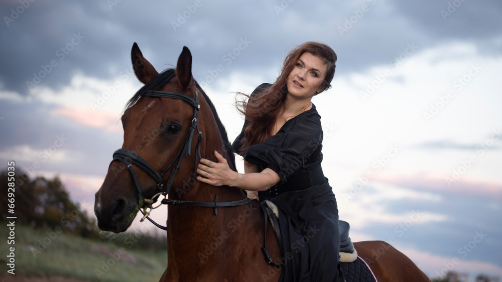 Obraz premium A beautiful young horsewoman in a black dress and with her hair down, riding a horse, portrait against the background of the evening sky, horseback riding in the open air