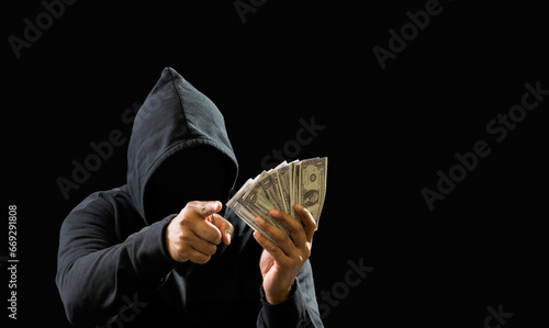 Portrait bandit man hacker one person wearing hood black shirt, sitting onchair and table thief hand holding money payment counting the amount obtained from hijacking or robbing, in dark background