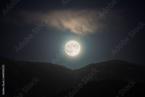 Full moon above mountains on cloudy sky, horizontal landscape of Earth satellite in full lunar phase