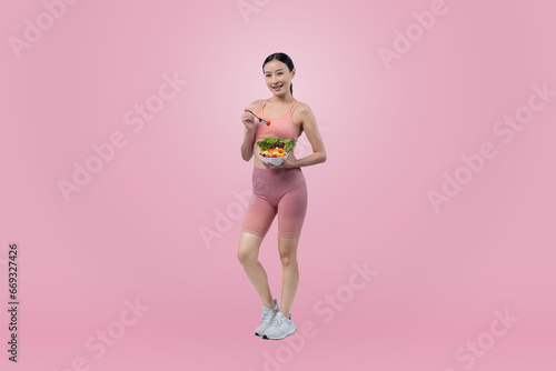 Fototapeta Naklejka Na Ścianę i Meble -  Young sporty Asian woman in sportswear holding salad bowl fill with vibrant of fruit and vegetable. Natural youthful and fit body lifestyle with balance nutrition on isolated background. Vigorous