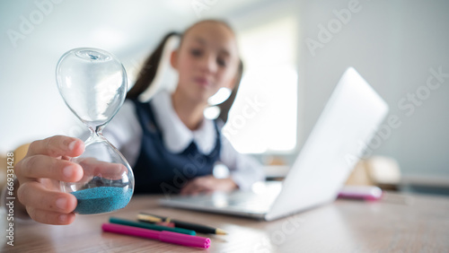 Caucasian girl sits at a desk at school and studies at a laptop. The schoolgirl turns over the hourglass.