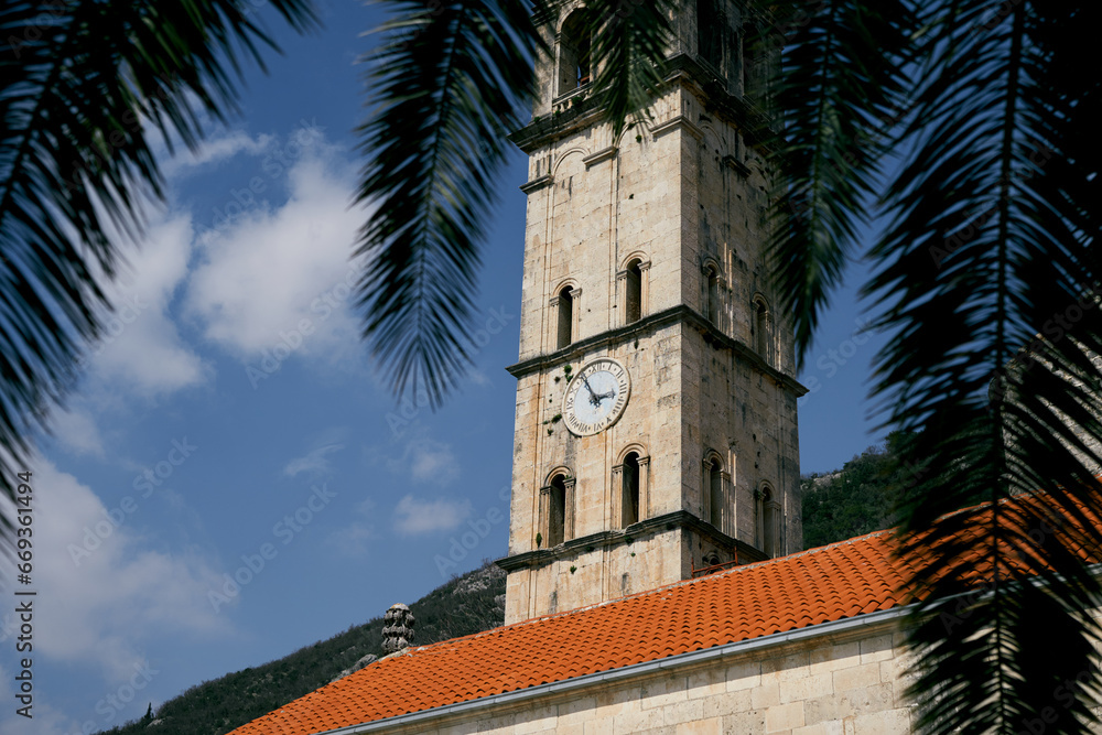 View through the palm branches to the bell tower with the clock of the Church of St. Nicholas. Perast, Montenegro
