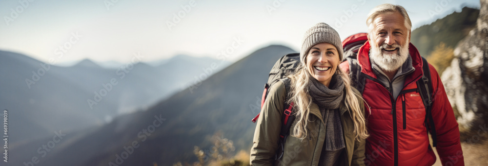 An elderly couple hiking, wearing winter clothes and standing against the backdrop of magnificent scenery - obrazy, fototapety, plakaty 