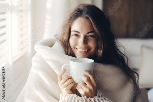 Young woman wrapped in a white blanket  holding a cup with hot drink trying to warm up in the cold apartment photo