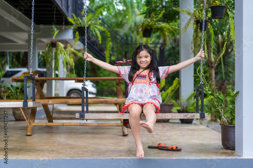 happy asian child or kid girl smiling sitting rest relax and playing on wooden swing with trees at home garden and family car on holiday and smile cheerful on green nature and wearing hilltribe outfit