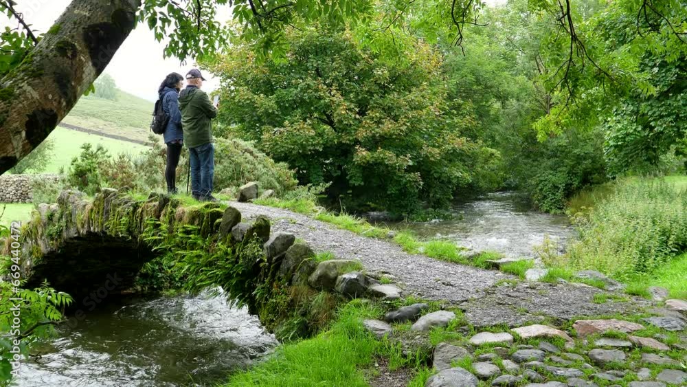 Mature couple standing on old small stone arch bridge over mountain stream talking and taking pictures in Lake District National Park, England, UK. Mature couple standing on old small stone arch bridge over mountain stream talking and taking pictures in Lake District National Park, England, UK.