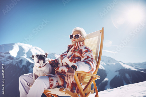 The older grandmother sits in a chair and enjoying in sun rays with her dog on a snowy mountain. Winter concept in ski trail in nature.