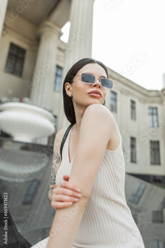 Stylish beautiful lady with vintage golden sunglasses in a fashion beige dress sits in the city. Pretty urban woman near a column building