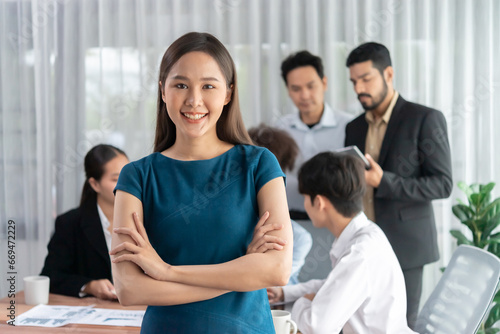 Young Asian businesswoman poses confidently with diverse coworkers in busy meeting room background. Multicultural team works together for business success. Office lady portrait. Concord © Summit Art Creations