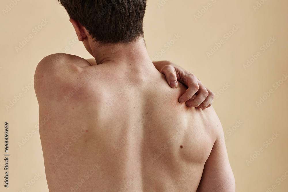 Close up of young man standing with bare back to camera and scratching shoulder, minimal Close up of young man standing with bare back to camera and scratching shoulder, minimal