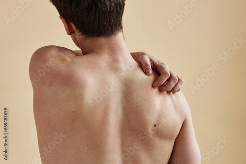 Close up of young man standing with bare back to camera and scratching shoulder, minimal