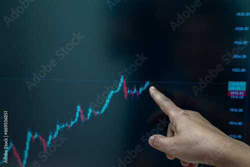 Hand of elderly woman points at the green bar chart of an investment or stock market on a computer screen.