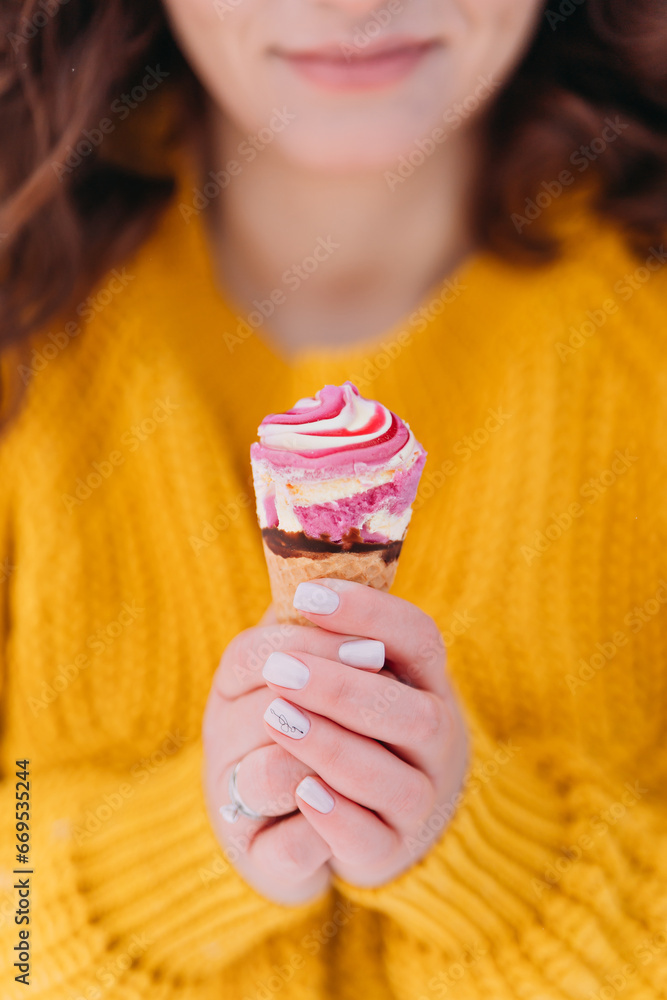 A pink ice cream cone in women's hands with a light manicure. The woman is wearing a bright yellow sweater. There are only hands in the frame