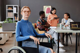 Portrait of young woman using wheelchair looking at camera in modern office with team in background, copy space