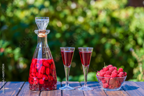 Homemade red raspberry brandy in two glasses and in a glass bottle on a wooden table in a summer garden, closeup