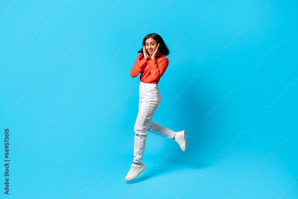 Excited Indian woman jumps and touching face against blue backdrop Excited Indian woman jumps and touching face against blue backdrop