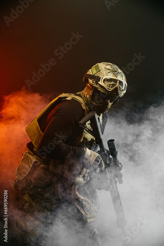 A soldier in combat gear with a weapon in his hands stands in battle formation among fire and smoke. The concept of military affairs. Computer games. The concept of war and combat.