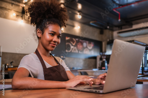 Cute young black female waitress barista using a laptop computer to check orders with happiness in the cafe at restaurant.