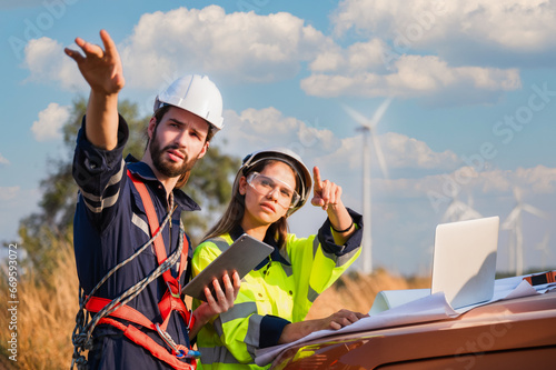 Two professional maintenance engineers in safety workwear using laptop and tablet checklist wind turbine on windmill construction farms.