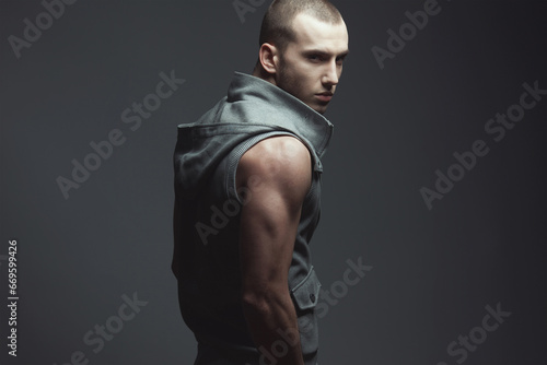 Male fashion concept. Portrait of handsome brutal young man with short hair, stubble on face wearing sleeveless jacket and posing over dark gray background. Street style. Copy-space. Studio shot photo